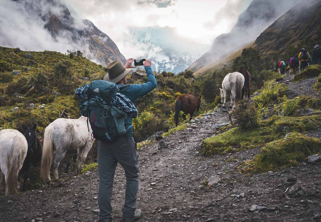 Turista tirando uma foto com o celular na Rota Salkantay, cercado por vegetação, neblina e cavalos