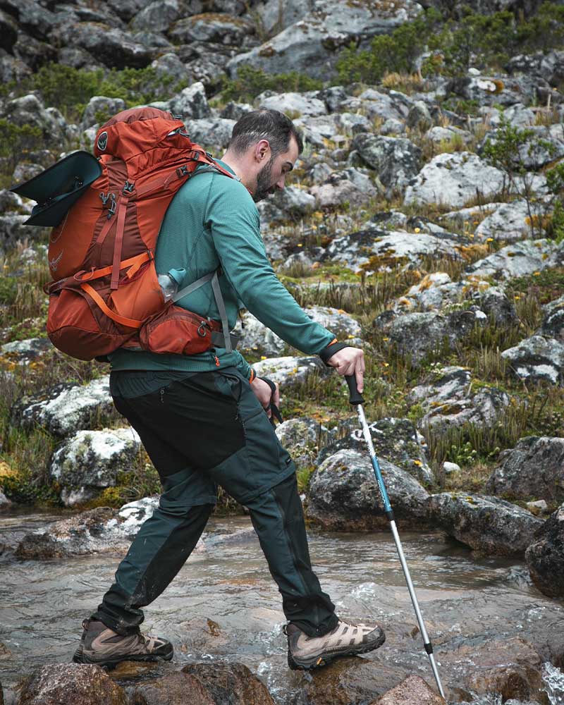 Caminhante atravessando um riacho durante a trilha de Salkantay, com bastões de trekking e uma mochila bem ajustada ao seu corpo