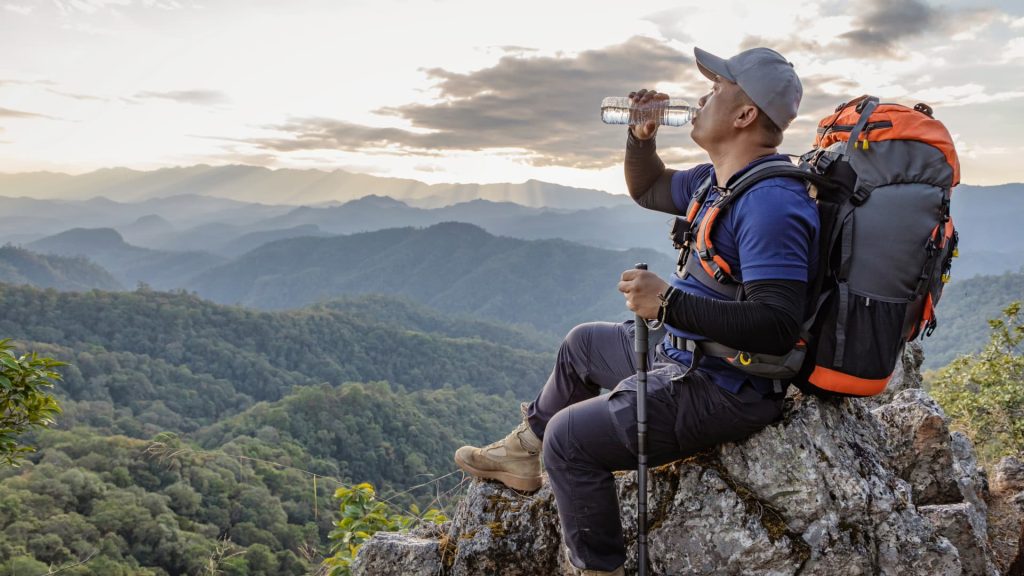 Um excursionista bebe água de uma garrafa enquanto está com dois amigos em uma trilha rochosa na montanha, com picos nevados.