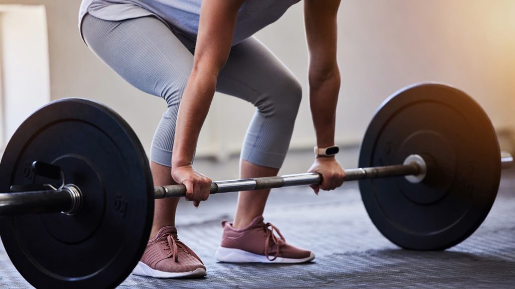 Primeiro plano de uma pessoa a segurar uma barra com pesos, a preparar-se para fazer um exercício de levantamento terra num ginásio.