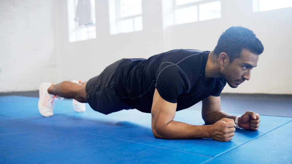 Uma pessoa em posição de prancha, apoiada nos antebraços, sobre um tapete azul, durante um treino de abdominais.