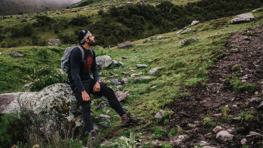 Uma pessoa cansada, com uma mochila às costas, a descansar sobre uma rocha durante um percurso exigente de montanha a grande altitude.