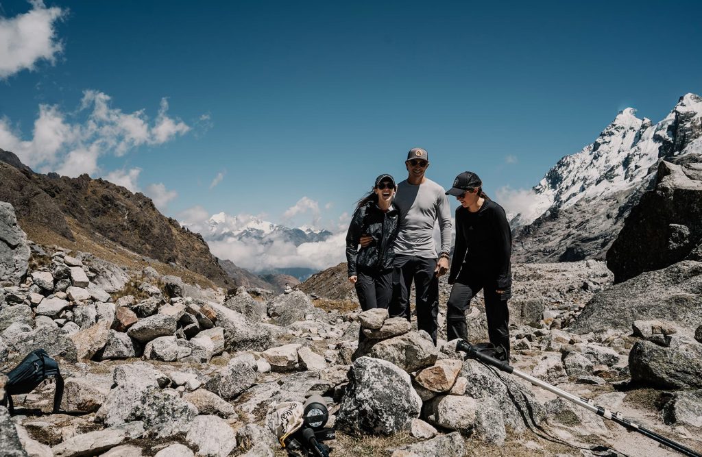 Três caminhantes sorrindo em uma trilha rochosa na montanha, com picos nevados ao fundo.